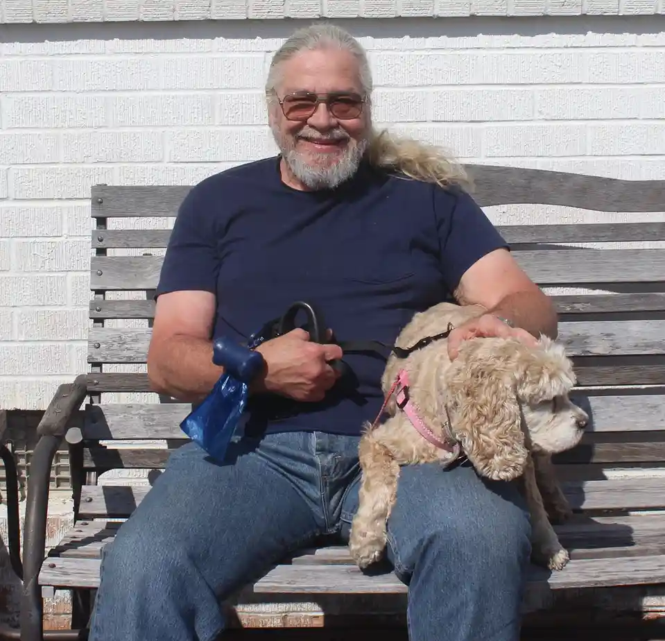 a smiling older man with a long gray ponytail and sunglasses sits on a wooden bench holding a small light-brown fluffy dog…