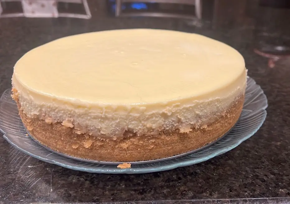 A whole, plain cheesecake with a golden-brown graham cracker crust sits on a clear glass plate atop a dark granite…