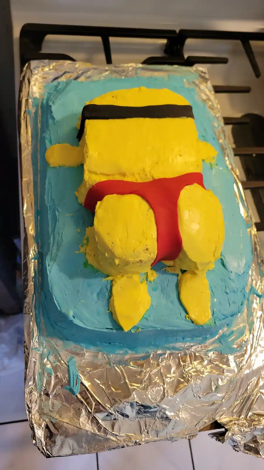A rectangular cake covered in blue frosting sits on aluminum foil, shaped and decorated to resemble a yellow Minion…