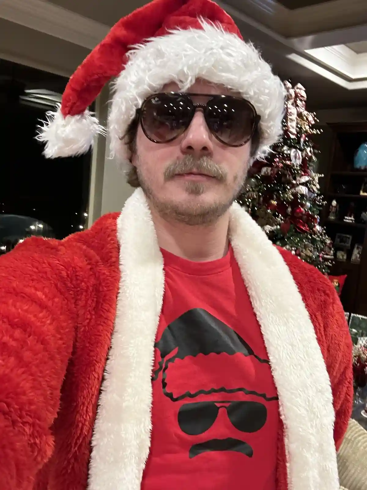A man wearing a red Santa hat, sunglasses, and a red fluffy robe over a red t-shirt with a graphic of a cool-looking Santa…