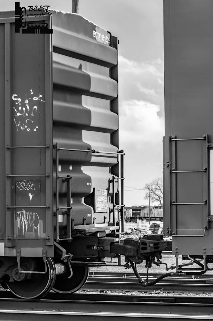 A black-and-white photograph of two freight train cars on tracks, one with visible graffiti. The image is overlaid with the…