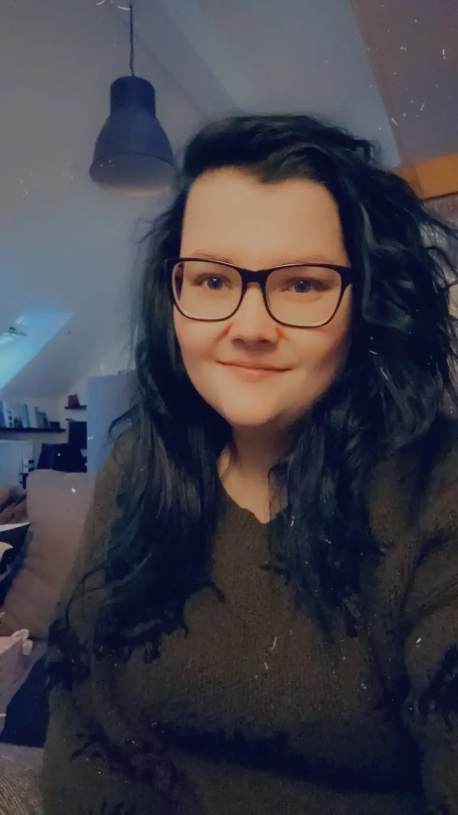 A person with dark, wavy hair and glasses smiles at the camera while wearing a dark sweater in a dimly lit room with a…