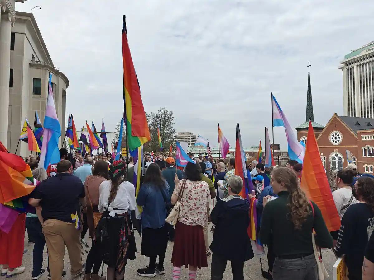 A crowd of people at an outdoor protest or rally, many holding rainbow and transgender pride flags, with a church and city…