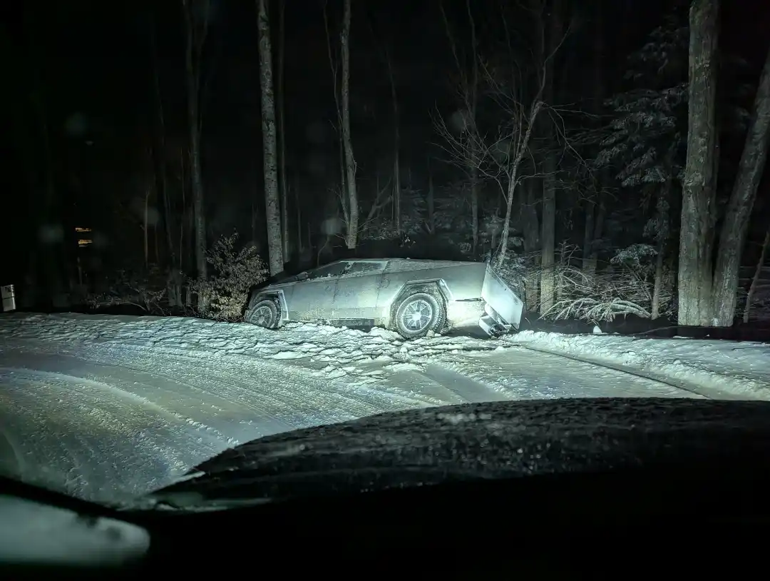 A nighttime photo taken from inside a vehicle shows a silver Tesla Cybertruck partially submerged in snow on the side of a…