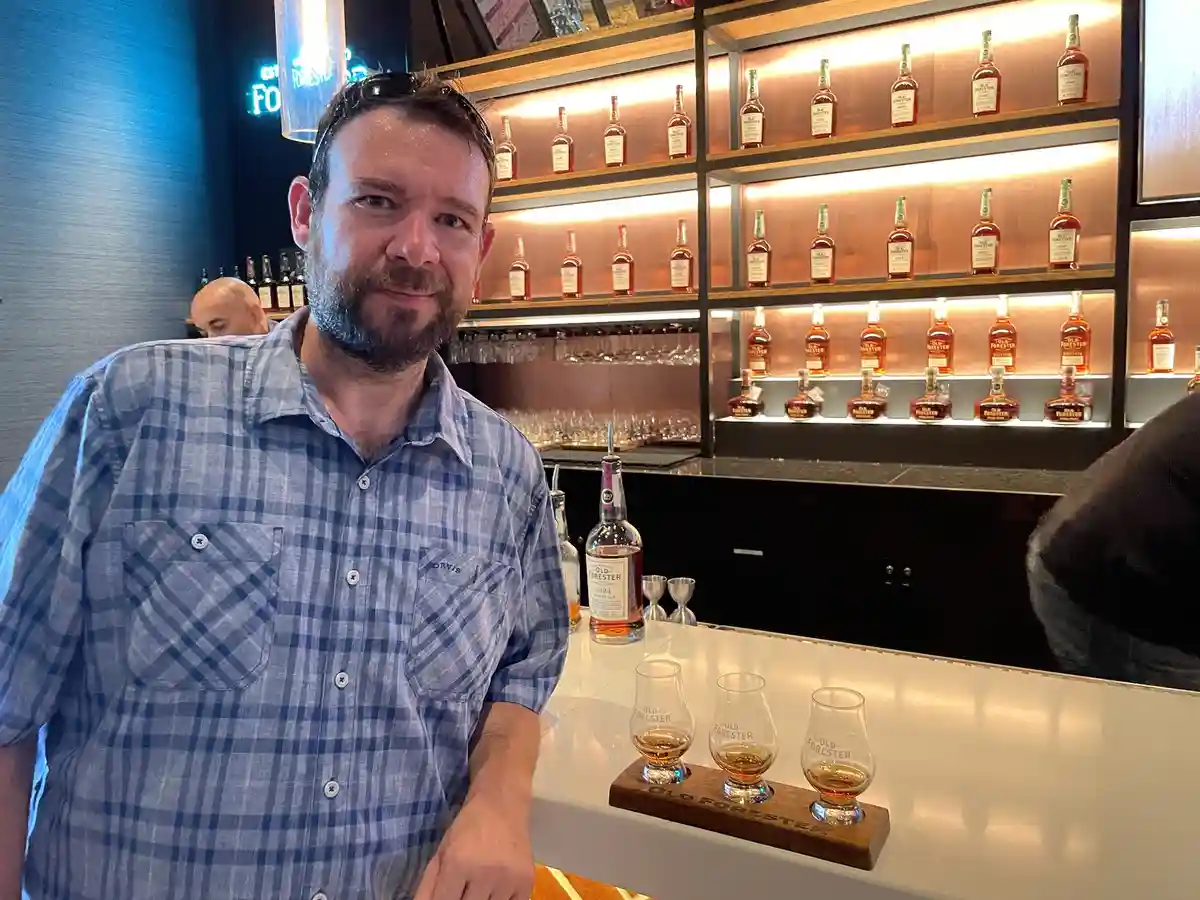 A man with a beard and sunglasses on his head stands at a bar with a blue plaid shirt, smiling slightly. In front of him on…