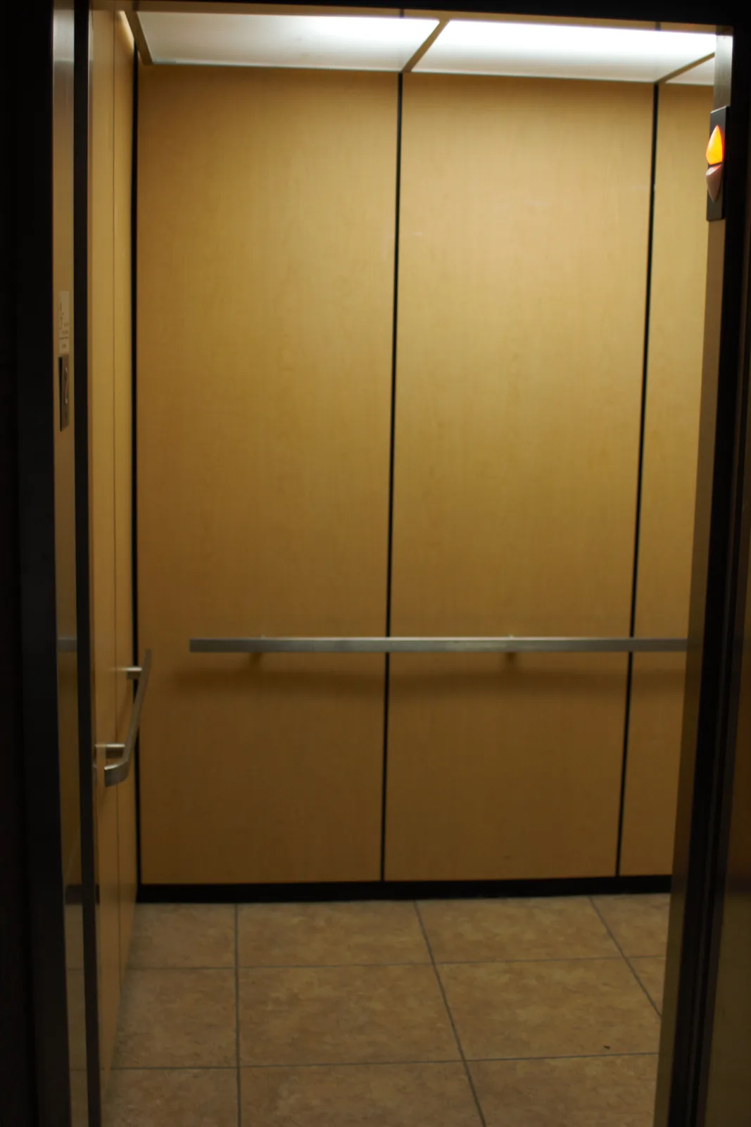 An empty elevator with light wood paneling, a metal grab bar, and tiled floor viewed from the doorway. The overlaid text…