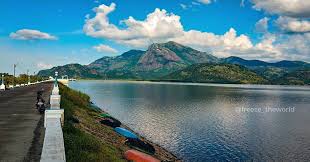 A scenic photo of a calm lake with mountains in the background and a paved path along the shore. A person is sitting on a…