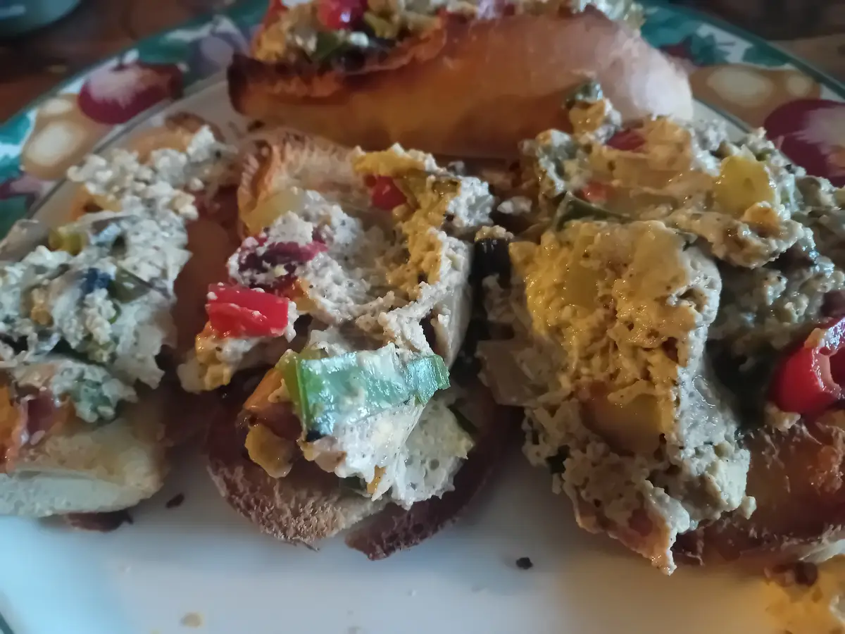 A close-up photo shows several pieces of bread topped with a creamy, chunky spread containing red and green peppers…
