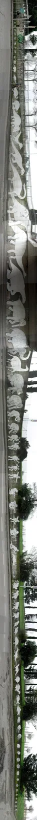 A long, vertical photograph shows a concrete wall with a series of white, stylized animal and human figures carved into it…
