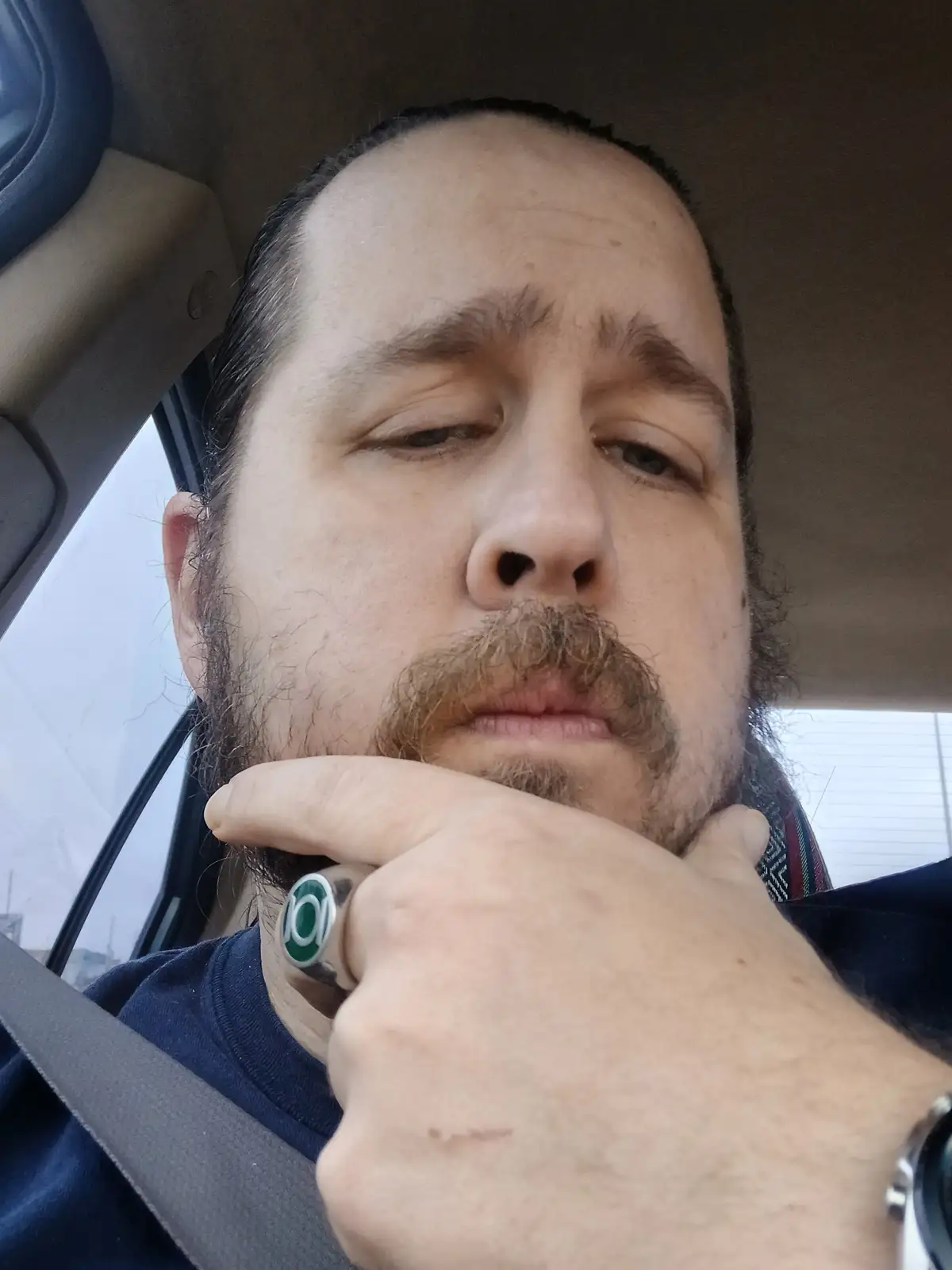 A man with a beard and mustache sits in a car, looking tired or bored, with his hand resting under his chin. He wears a…