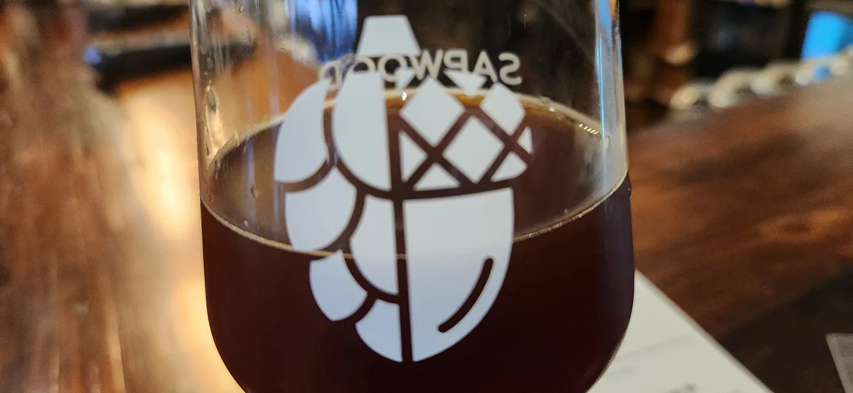 A close-up photo of a glass of dark beer with a white logo on it that reads 'sabwood' and features a stylized tree design…