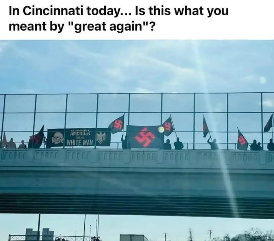 A photo taken from a car shows silhouettes of people standing on a bridge railing, waving multiple black flags with red…