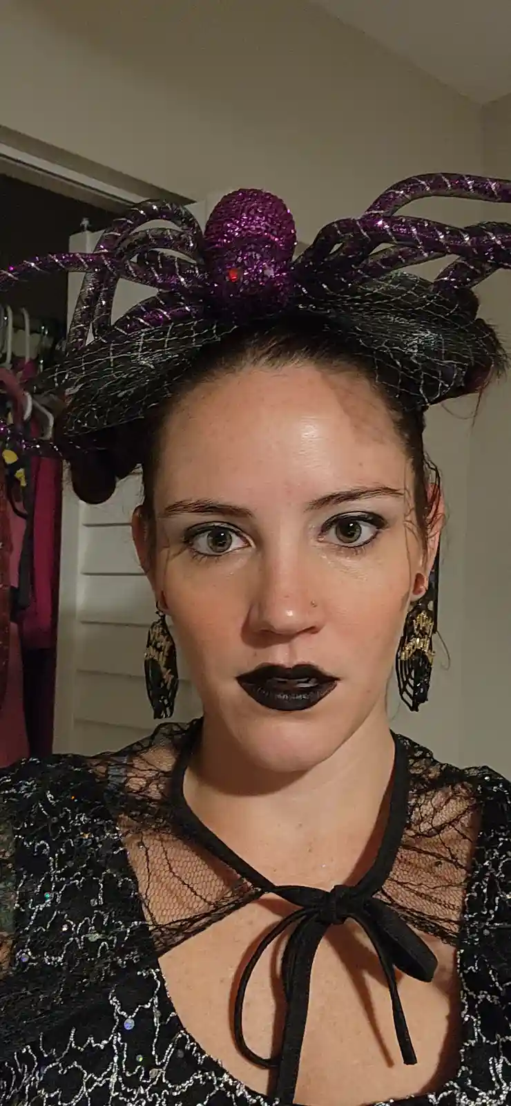 A woman with dark lipstick and dramatic eye makeup wears a large, glittery purple spider headpiece with black webbing…