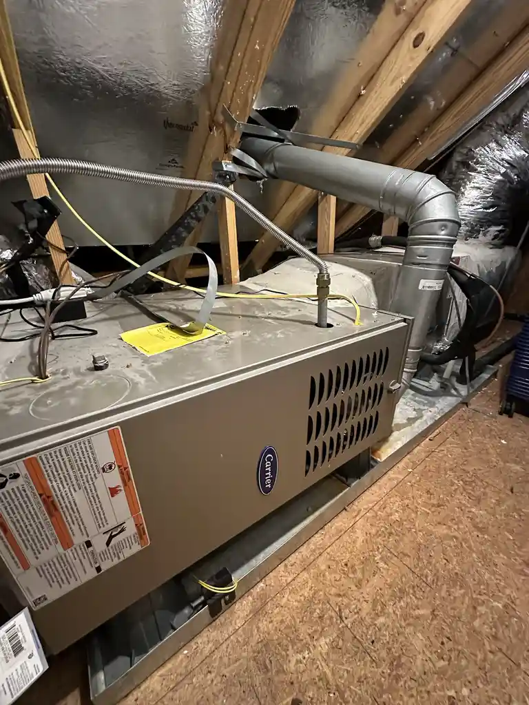 A close-up photo of a Carrier furnace installed in an attic space, surrounded by exposed wooden beams, ductwork, and…