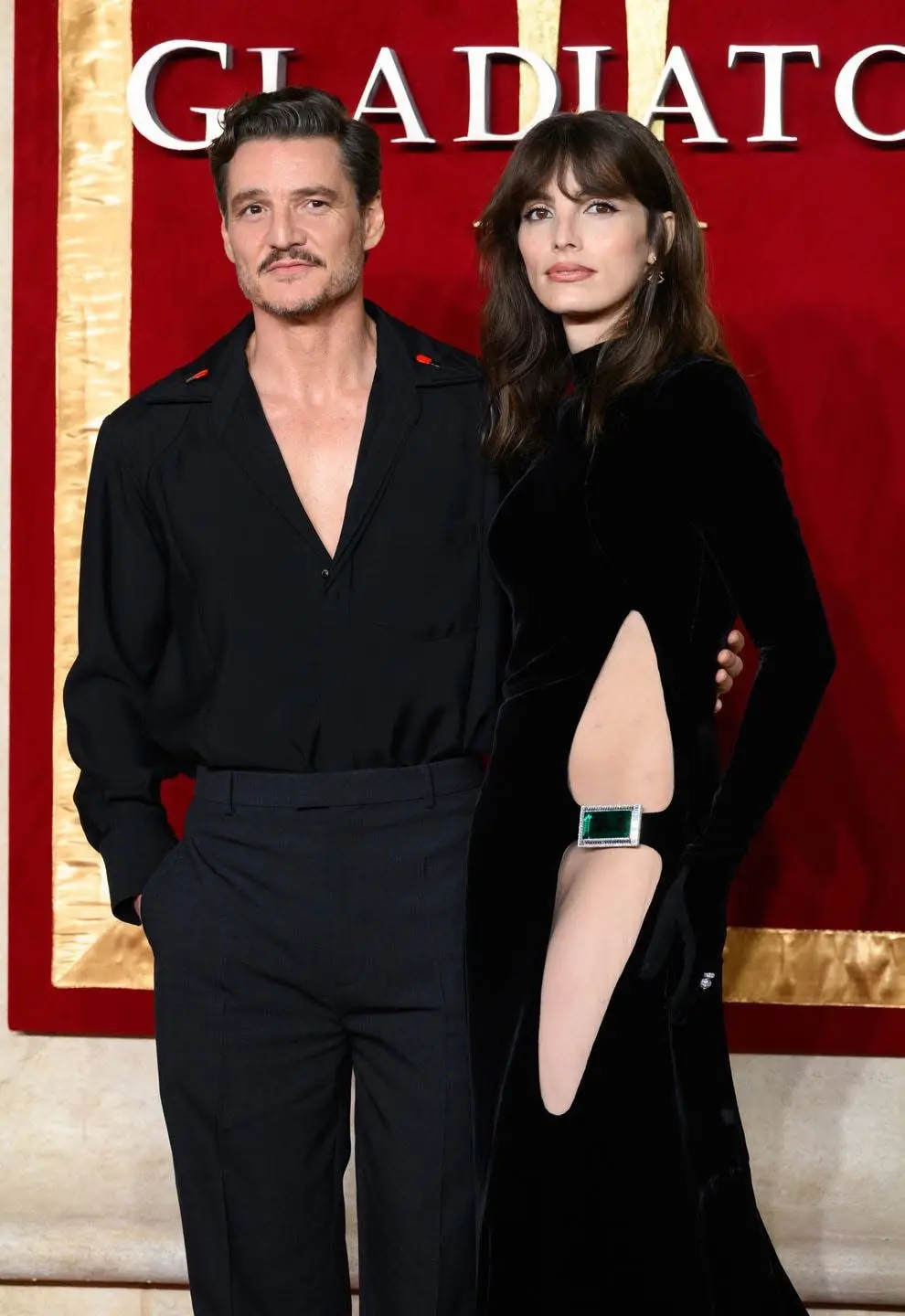 A man and woman pose together at a red carpet event in front of a backdrop with the word 'GLADIATOR' in large white letters…