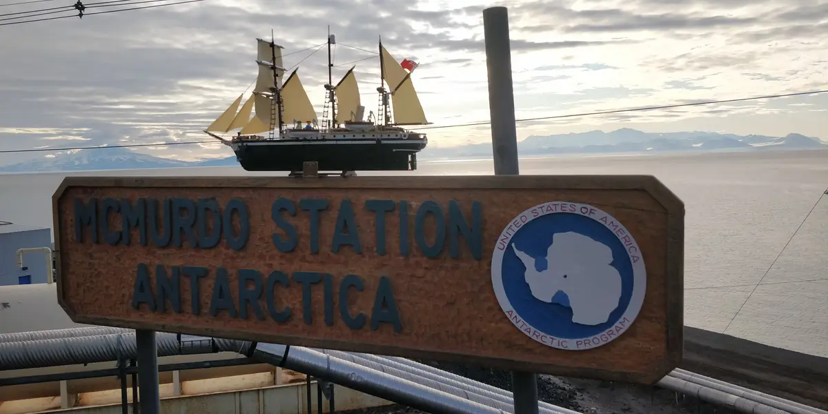 A wooden sign reading 'MCMURDO STATION ANTARCTICA' with a USA Antarctic Program logo, topped by a model of a sailing ship…
