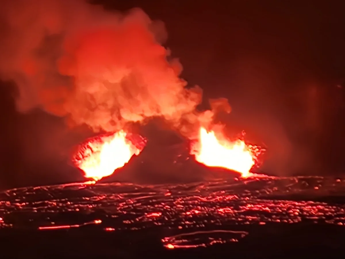 A nighttime volcanic eruption shows two glowing lava fountains erupting from a lava field, sending plumes of red-orange…