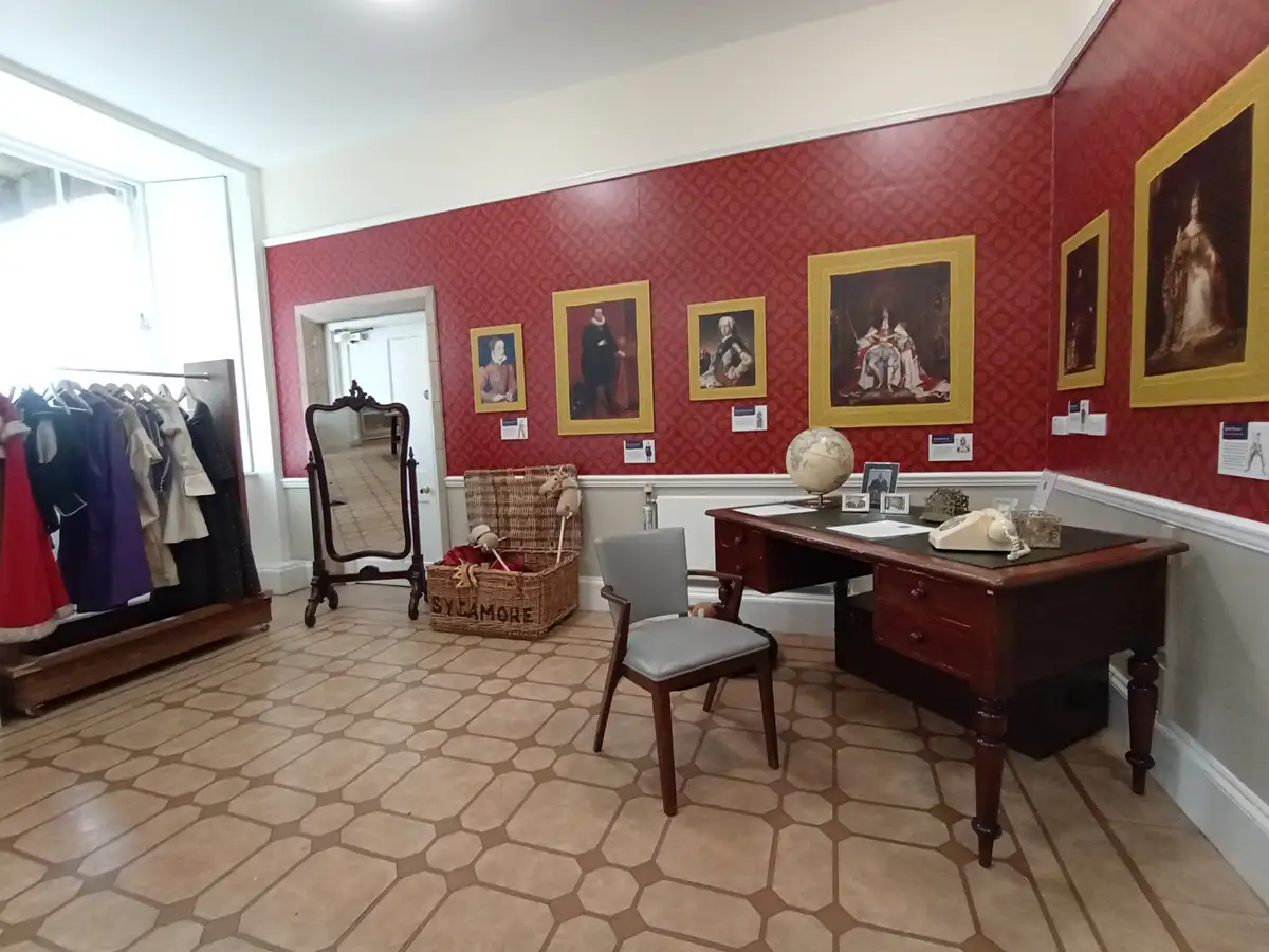 A room with red patterned wallpaper, a wooden desk with a globe and rotary phone, a wicker basket labeled 'SYCAMORE'…
