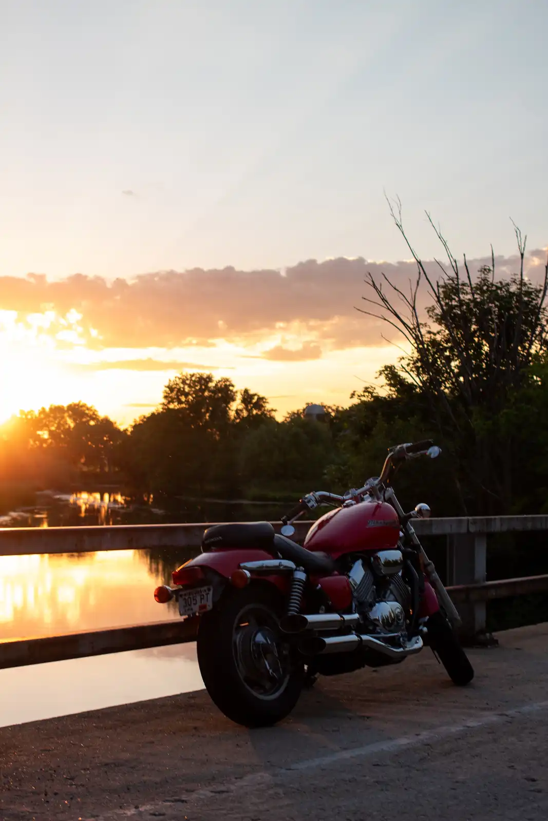 A red Honda motorcycle is parked on a bridge railing overlooking a river at sunset, with warm orange and pink clouds in the…