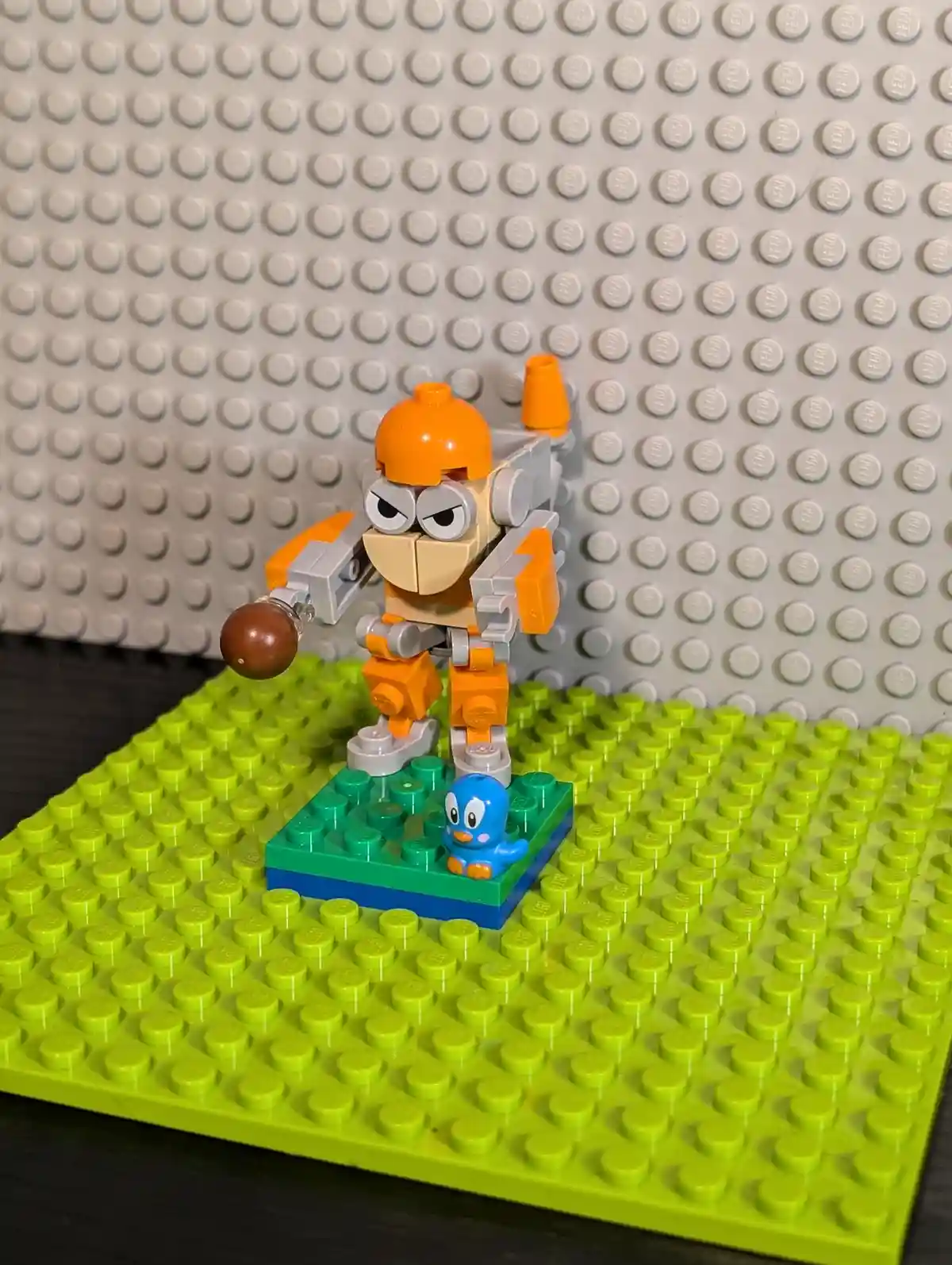 A Lego minifigure with an orange helmet, angry eyes, and a brown ball in its hand stands on a green baseplate. A small blue…