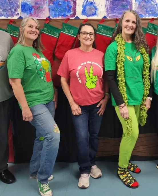 Three women pose in front of decorated Christmas stockings. The woman on the left wears a green shirt with a Grinch graphic…