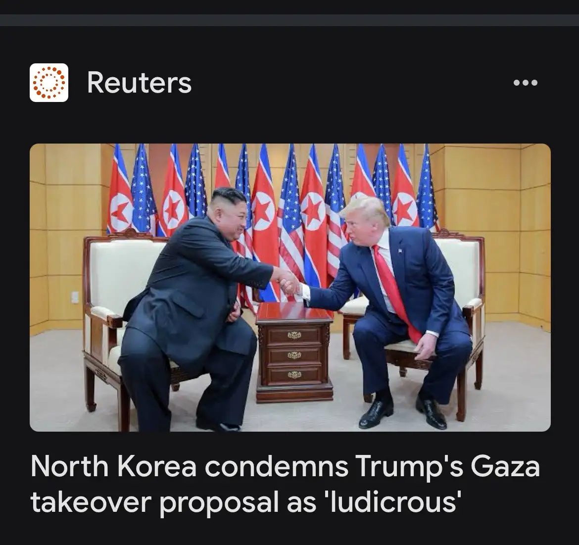 Two men in suits shake hands across a small table while seated in chairs, with alternating North Korean and American flags…