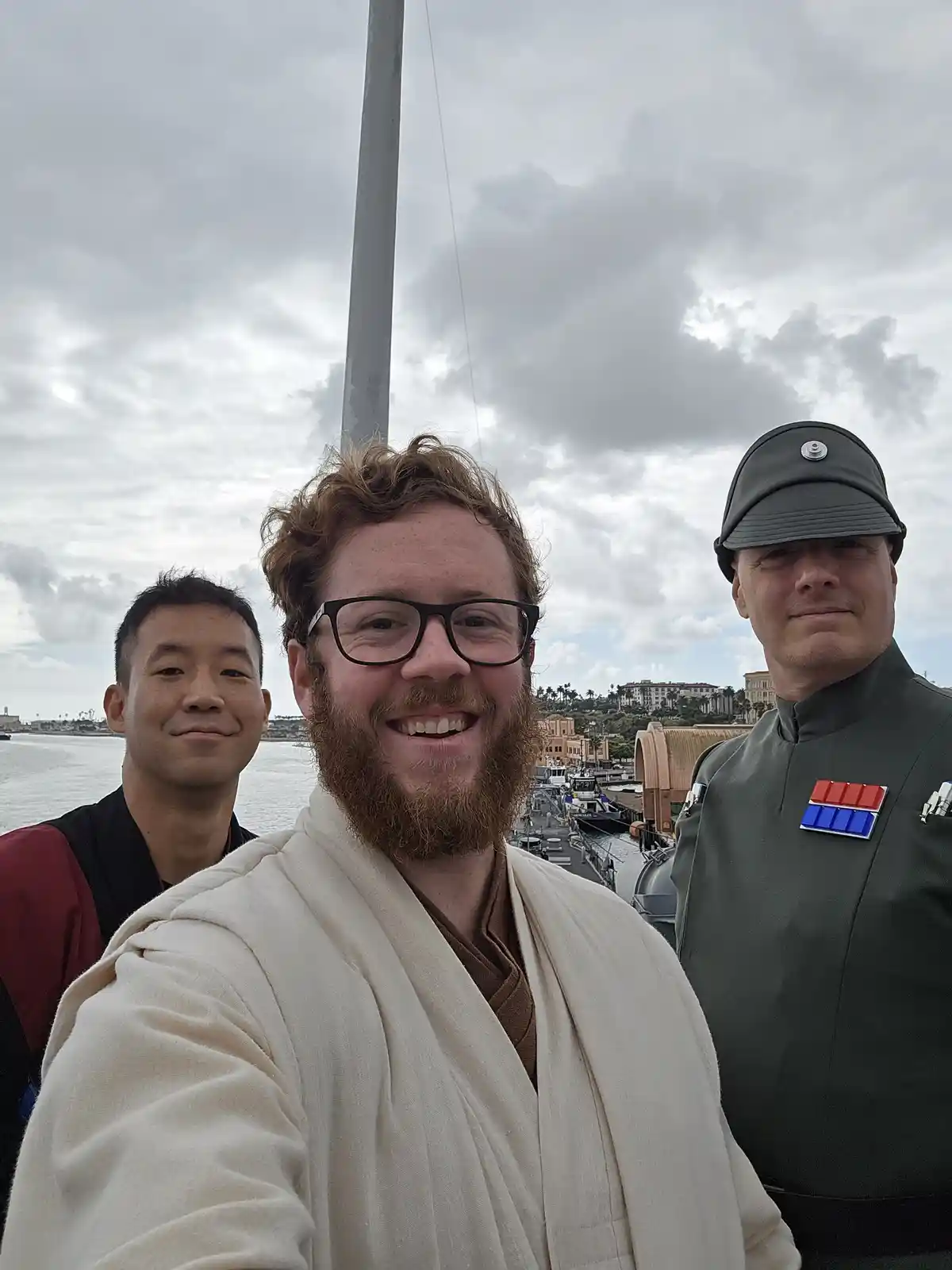 Three men pose for a selfie on a boat deck with a cloudy sky and harbor in the background. The man in the center, wearing…