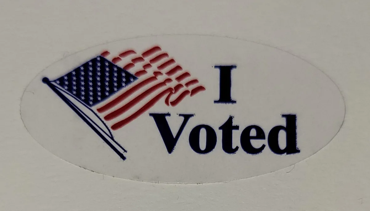 A close-up of a white oval sticker featuring a stylized American flag waving to the left, with the text 'I Voted' in bold…