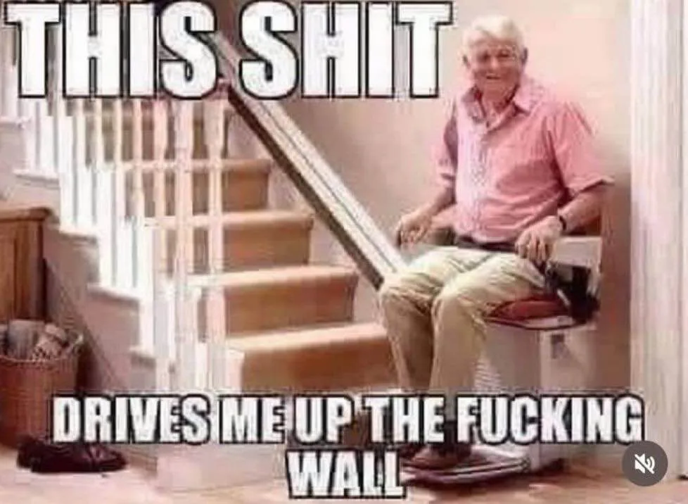 An older man in a pink shirt and khaki pants sits on a stairlift that is positioned at the bottom of a wooden staircase. The…