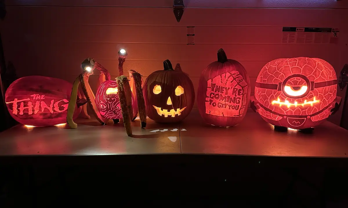 Five intricately carved and lit pumpkins sit on a table under red lighting. From left to right: one carved with 'THE THING'…