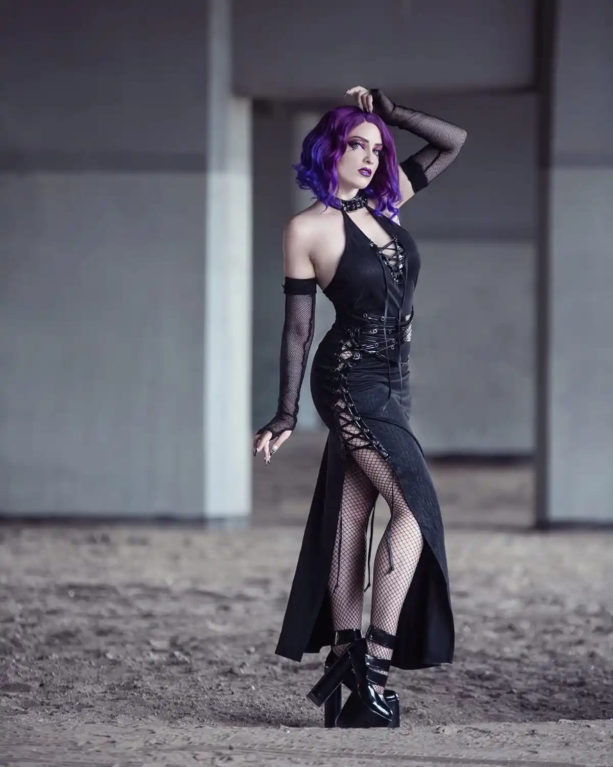 A woman with purple hair poses in a black, lace-up, fishnet outfit with high heels in a concrete structure. She has one hand…