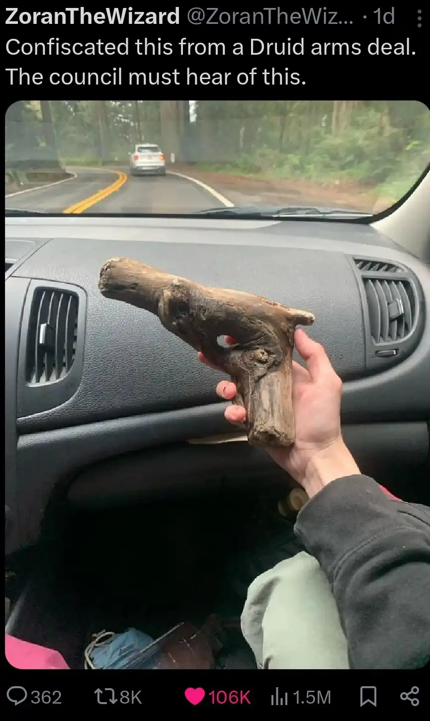 A hand holds a piece of driftwood shaped like a handgun inside a car, with the dashboard and a road visible through the…