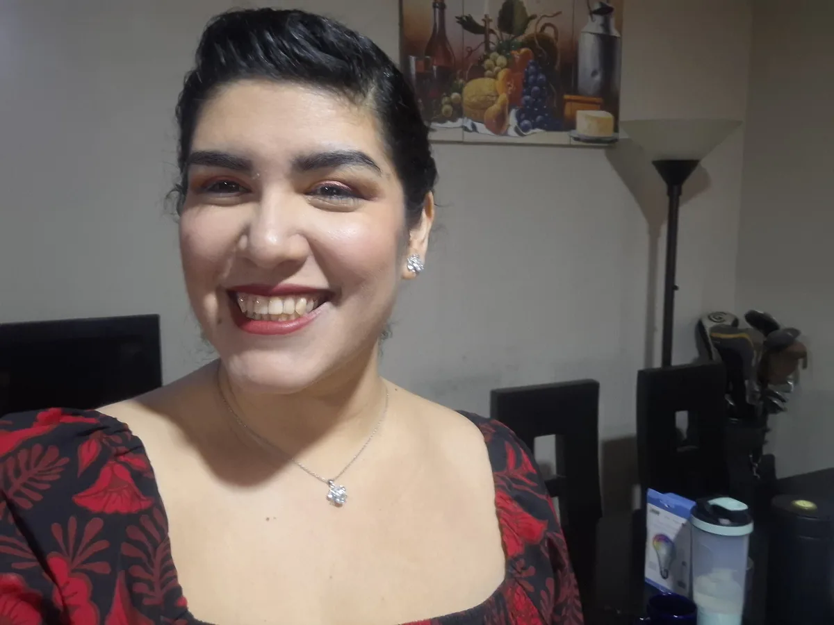 A woman with dark hair styled up, wearing a red and black floral top, a silver necklace with a flower pendant, and sparkly…