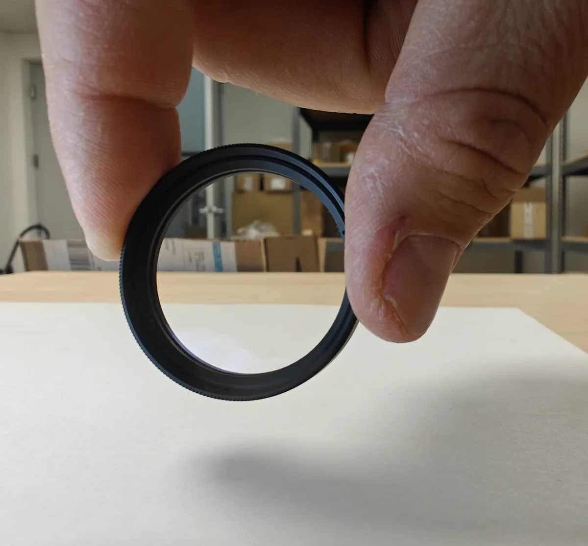 A hand holds a black, circular camera lens filter ring against a white surface, with a blurred background of shelves and…