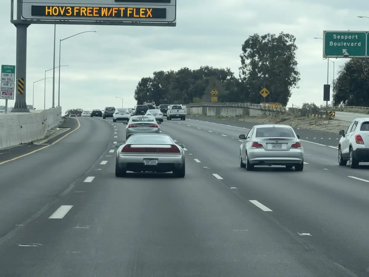 A highway scene with multiple cars in traffic under an electronic sign that reads 'HOV3 FREE W/FT FLEX'. A green road sign…