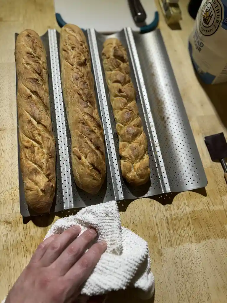 Three freshly baked loaves of bread, two with slashes and one braided, sit on a perforated metal baking tray on a wooden…