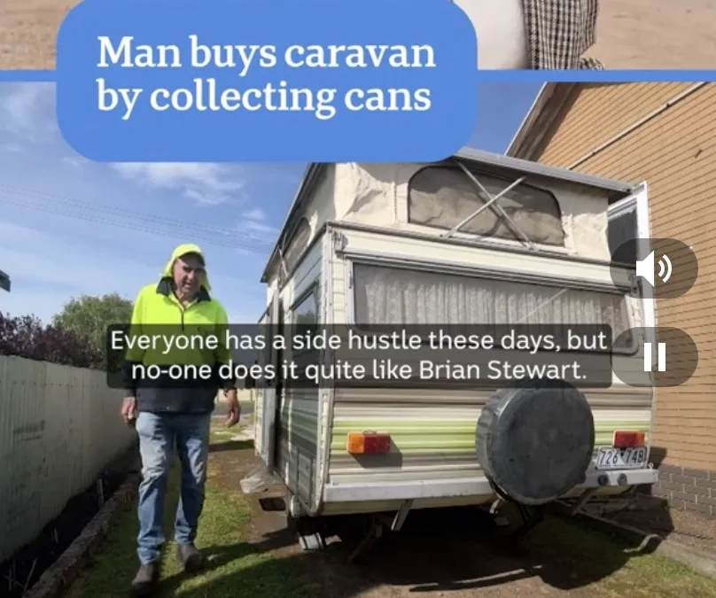 A man in a bright yellow hoodie and jeans walks toward the camera beside a beige and green caravan parked in a yard. A blue…
