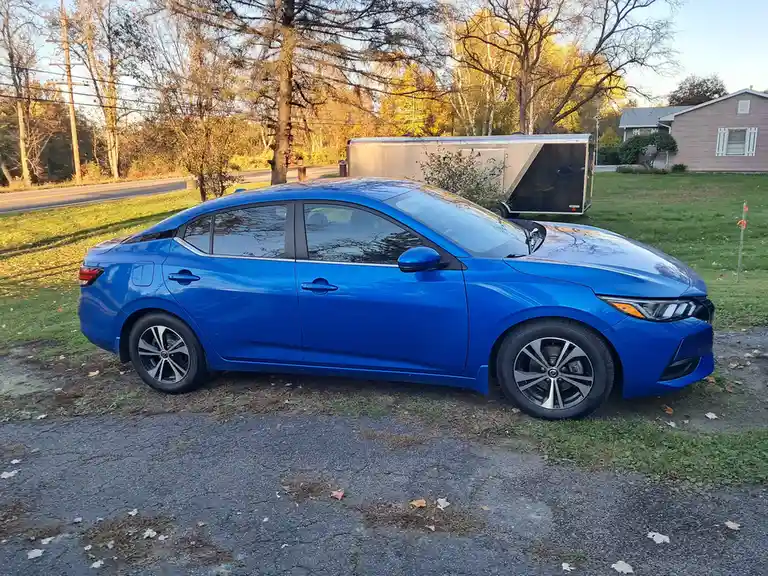 A bright blue sedan is parked on a gravel driveway in front of a house and a trailer, with autumn trees and a road in the…