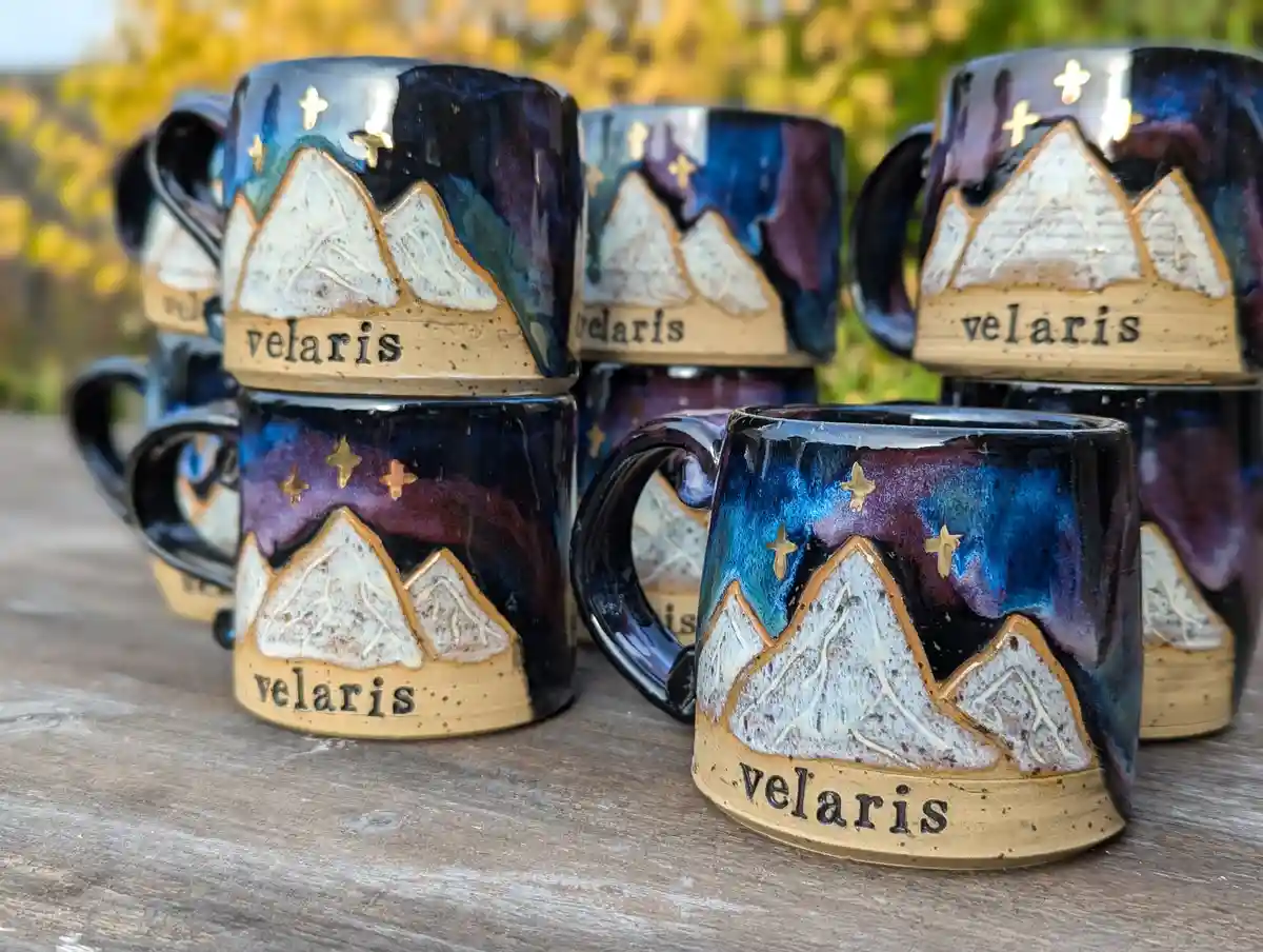Several handmade ceramic mugs with a mountain and star design are stacked and arranged on a wooden surface. Each mug…