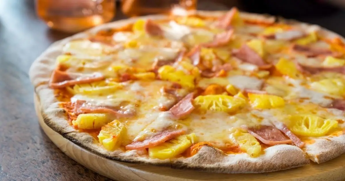 A close-up photo of a Hawaiian pizza with melted cheese, ham slices, and pineapple chunks on a wooden serving board. The…