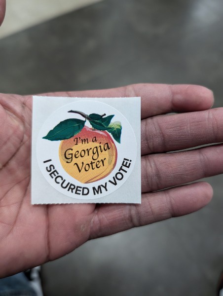 A person holds a circular sticker in their open palm. The sticker features a peach illustration with green leaves and the…