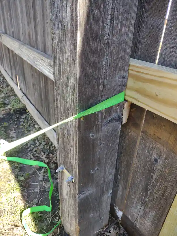A bright green strap is tied around a weathered wooden fence post, connecting it to a new light-colored wooden beam. The…
