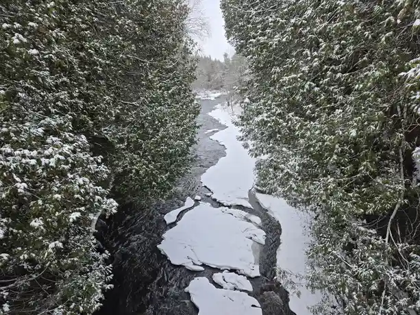 A snowy winter scene shows a river flowing through a forest, with snow-covered evergreen trees on both sides and large…