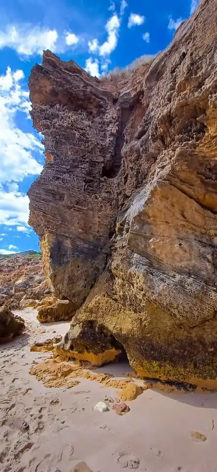 A vertical photo of a sandy beach with towering, layered rock formations under a bright blue sky with white clouds. The…