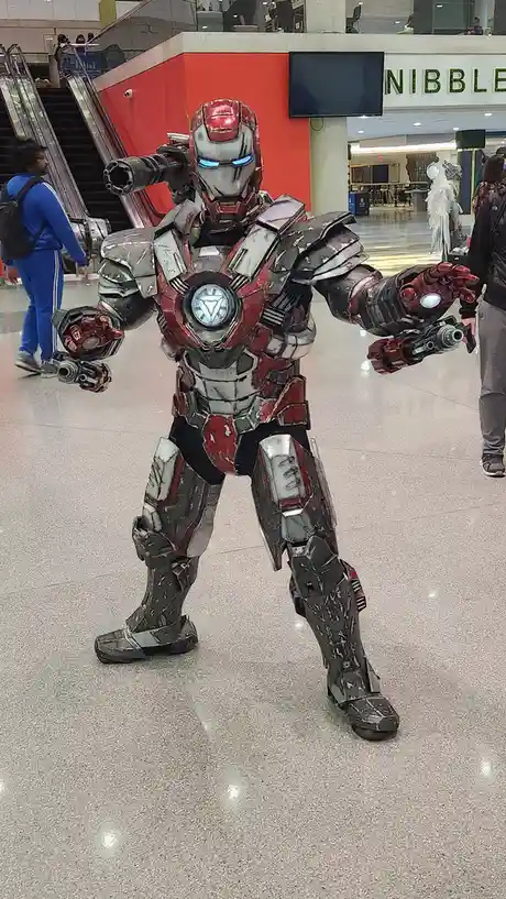 A person in a heavily weathered, red-and-gray Iron Man suit with glowing blue eyes and multiple mounted weapons stands in a…