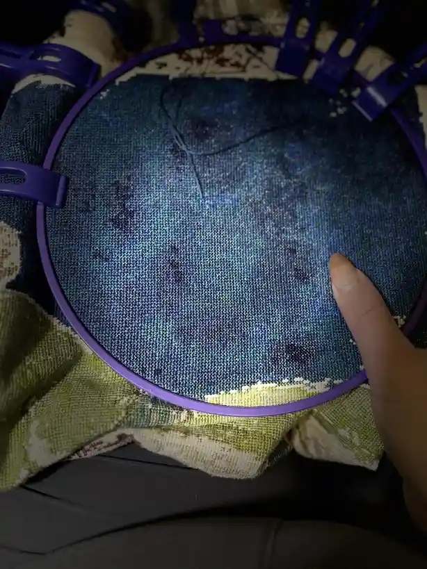 A close-up photo shows a person's finger resting on a purple embroidery hoop holding a blue fabric with a starry night…