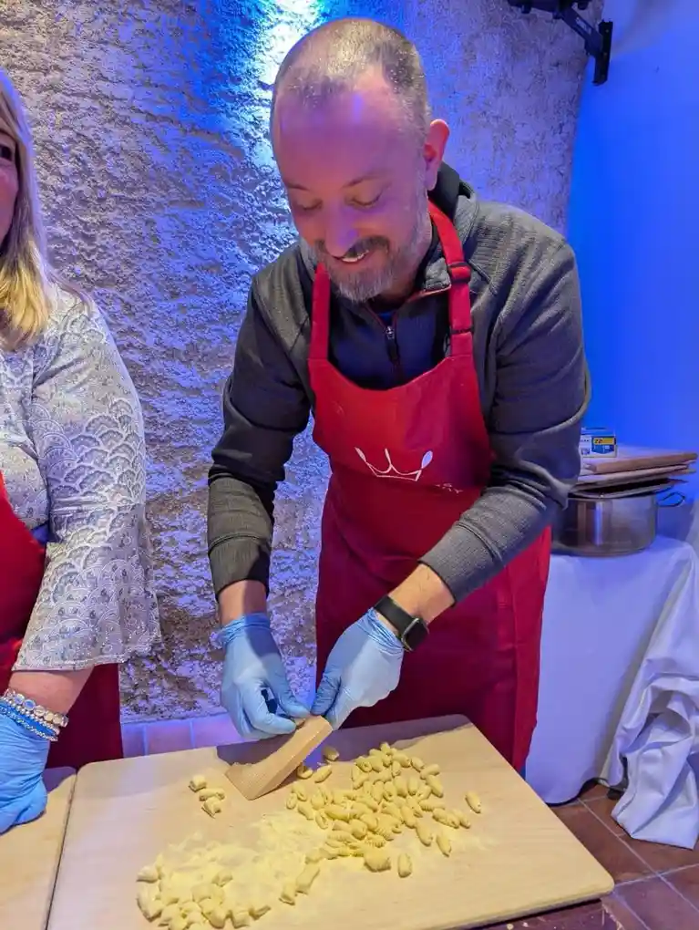 A man in a red apron and blue gloves smiles while shaping pasta on a wooden board, with a woman partially visible beside…