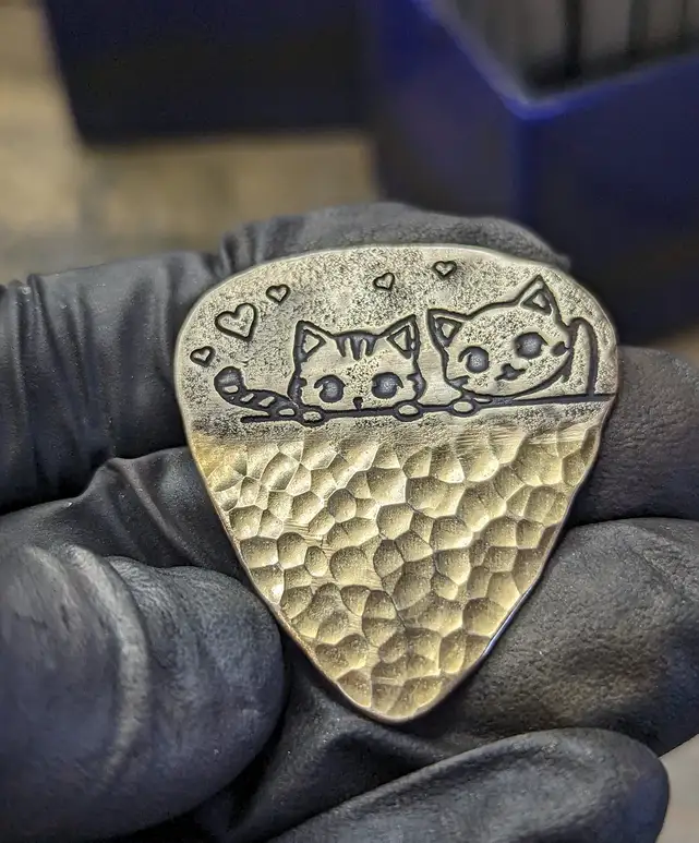 A hand wearing a black glove holds a custom-made metal guitar pick. The pick features an engraved design of two cute cartoon…