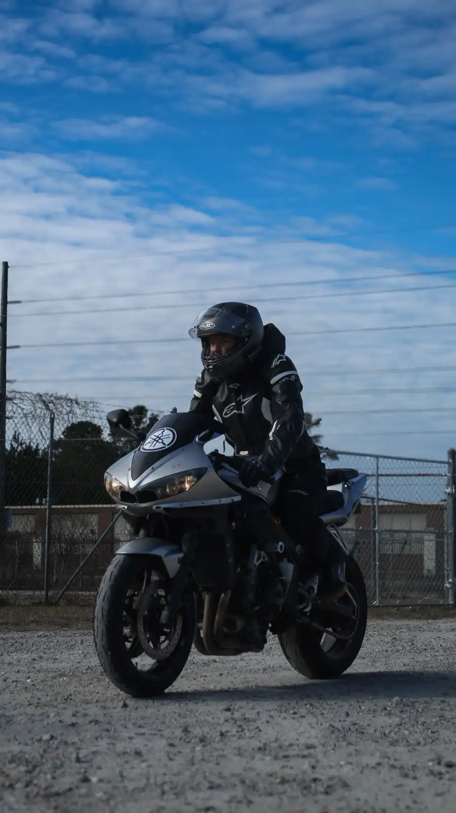 A person in full black motorcycle gear, including a helmet, sits on a silver Yamaha sportbike parked on gravel. The…