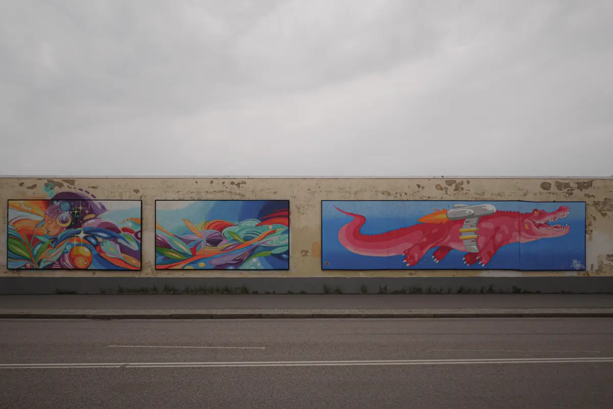Three colorful murals are painted on a concrete wall beside a road under an overcast sky. The left mural depicts a stylized…